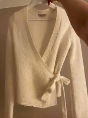 Vit omlottkofta från Gina tricot - Mysig vit omlottkofta från Gina tricot  med knyt i sidan och djup v-ringning. Koftan är stickad och har långa ärmar samt ribbade kanter vid hals och nederkant. Perfekt för lager-på-lager och chill dagar.