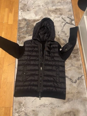 Svart Moncler hoodie med huva - Säljer en svart hoodie från Moncler med quiltad framsida och stickade ärmar. Tröjan har huva, dragkedja framtill och en ficka med dragkedja på ärmen. Perfekt för höst och vinter, riktigt snygg och bekväm med exklusiv känsla.