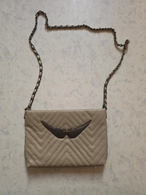 Beige quiltad axelväska med kedja - Snygg beige axelväska med quiltad yta och coolt metallspänne i form av vingar på framsidan. Väskan har en kedja med insydda detaljer och flera fack inuti, inklusive ett med dragkedja. Perfekt för dig som gillar stilrena accessoarer med en twist.