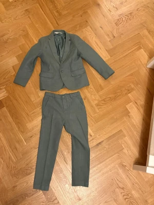 Grön kostym med kavaj och byxa strl 128 - Snygg grön kostym med klassisk kavaj och matchande byxa. Kavajen har två framfickor, knappstängning och stilren krage. Byxorna har raka ben och fickor på sidorna. Materialet känns lätt och passar perfekt för dig som vill ha en clean look.