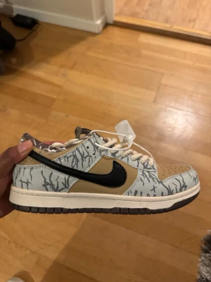 Nike Dunk Low med beige och svart - Nike Dunk Low sneakers med beige och grå paneler, svart swoosh och vitt snörning. Skorna har ett unikt grått mönster på ovandelen och brunt foder med grafiskt tryck. Perforerad tå och platt sula för skön komfort. Perfekt för dig som gillar streetwear och snygga detaljer.