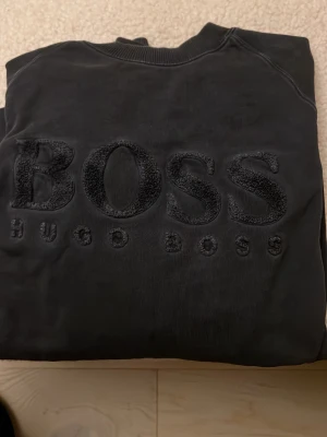 Svart sweatshirt från Hugo Boss - Säljer en svart sweatshirt från Hugo Boss med stort, broderat BOSS-tryck på bröstet. Tröjan har rund hals och ribbade muddar. Perfekt för dig som gillar stilrena och klassiska märkesplagg.