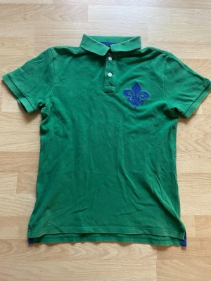 Grön pikétröja från Morris - Snygg grön pikétröja från Morris Stockholm med blå broderad lilja på bröstet. Klassisk krage, korta ärmar och tre vita knappar framtill. Tillverkad i bomull som känns skön mot huden. Perfekt för dig som gillar stilrena detaljer och vill sticka ut med färg. Sitter som S