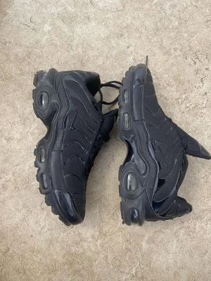 Nike Air Max Plus svarta sneakers - Nike Air Max Plus sneakers i helsvart färg med synliga Air-enheter i sulan. Skorna har en mesh- och syntetöverdel med vågiga detaljer, snörning och rund tå. Snyggt Nike Air-märke på plösen och robust platt sula för extra komfort.