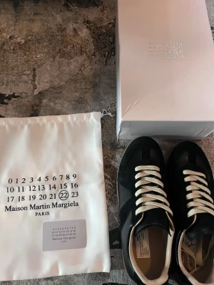 Maison Margiela skor - Maison Margiela sneakers i svart med kontrasterande vita snören och beige insida. Snygg retrostil med gummisula i ljusbrun och klassisk logotyp på sulan. Materialmix av skinn och mocka, levereras med originalpåse och låda.