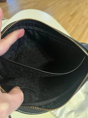 Svart axelväska från Michael Kors - Säljer en stilren svart axelväska från Michael Kors i äkta skinn med guldfärgade metalldetaljer och kedja. Väskan har ett rymligt fack med innerficka och dragkedja. Perfekt storlek för det viktigaste och snygg att bära över axeln.