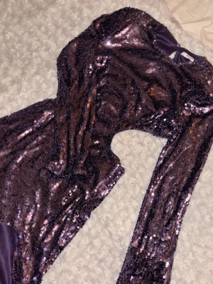 Lila paljettklänning från NA-KD - Säljer en glittrig lila kortklänning från NA-KD med långa ärmar. Klänningen är täckt av paljetter och har en knapp i nacken för stängning. Klänningen har en öppen rygg och öppna sidor. Jag har använt den endast en gång, så sicket är väldigt bra!