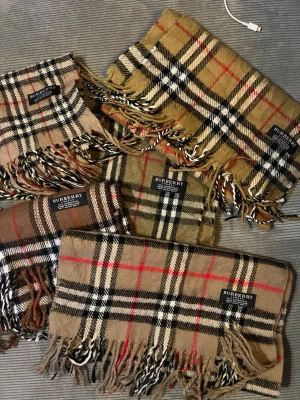 Burberry Scarf - Säljer en klassisk rutig scarf från Burberry i 100% lammull. Västen har det ikoniska rutmönstret i beige, brunt, svart, vitt och röda linjer, samt fransar längs nederkanten. Perfekt för dig som vill ha en tidlös och snygg look med premiumkänsla.