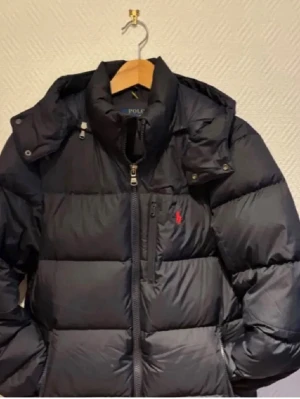 Svart pufferjacka från Polo Ralph Lauren - Svart pufferjacka från Polo Ralph Lauren med hög krage, dragkedja och RL-detaljer på knappar och zip. Jackan har snörning vid huvan och ett rött Polo-logo broderat på bröstet. Perfekt för kalla dagar och har en schysst, modern look.