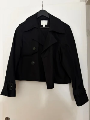 Svart croppad trenchcoat från H&M - Stilren svart croppad trenchcoat från H&M med bred krage, stora knappar och snygga spänndetaljer vid ärmsluten. Jackan har en loose passform och är tillverkad i ett slitstarkt polyestermaterial. 