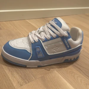Louis Vuitton blåvita sneakers - Snygga Louis Vuitton sneakers i vitt och ljusblått med klassiska LV-loggor på sidan, tungan och hälen. Skorna har snörning, platt sula och är tillverkade i skinn och syntet. Perfekta för dig som vill sticka ut med exklusiv streetstyle.