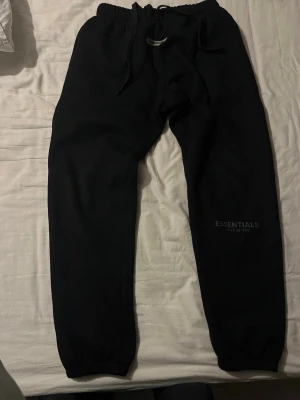 Svarta mjukisbyxor Essentials M - Svarta mjukisbyxor från Essentials Fear of God i storlek M. Byxorna har elastisk midja med snörning och ribbade benslut. Diskret Essentials-logga på benet och mjukt material som är skönt att ha på sig. 