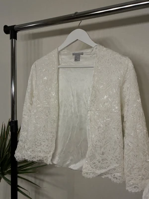 Vit spetskofta med blommigt mönster - Vit oanvänd kofta från H&M med blommigt spetsmönster och glittriga detaljer. Koftan har långärmad design och är öppen framtill. Perfekt att slänga över en topp för extra stilpoäng.