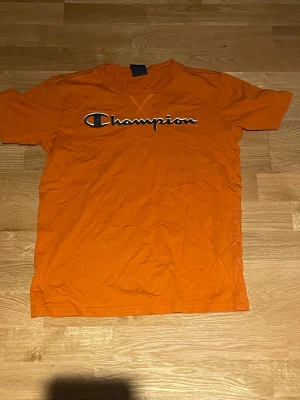 Orange Champion t-shirt med tryck - Säljer en orange t-shirt från Champion med stort logotryck över bröstet. Klassisk rund hals och korta ärmar. Perfekt för dig som gillar sportiga och färgstarka plagg. Materialet är mjuk bomull som känns skönt mot huden.