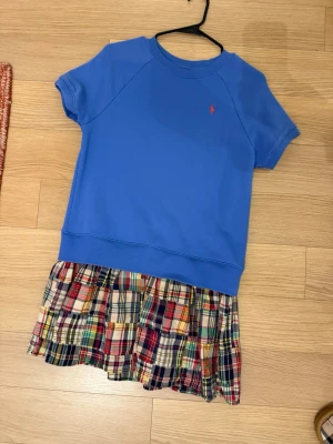 Blå kortklänning från Polo Ralph Lauren - Snygg kortklänning från Polo Ralph Lauren med blå överdel i bomull och korta ärmar. Kjoldelen har ett färgglatt rutigt mönster i flera nyanser som ger en cool kontrast. Perfekt för dig som gillar sportig och lekfull stil.