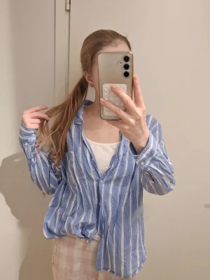 Blåvit randig skjorta - Säljer en blå skjorta med vita ränder. Skjortan har klassisk krage, långa ärmar och knäppning framtill. Tillverkad i mjuk bomull, perfekt för en chill och avslappnad stil.