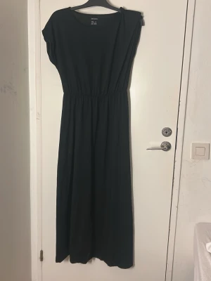Svart långklänning från Esmara - Svart långklänning från Esmara i storlek M. Klänningen har rund halsringning, korta ärmar och ett elastiskt midjeband som ger en snygg siluett. Tillverkad i mjukt bomullsmaterial som känns skönt mot huden. Enkel och stilren design som är lätt att matcha.
