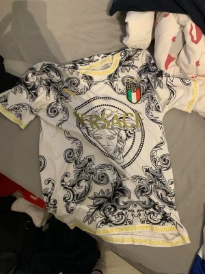 Fotbollströja - Säljer en riktigt unik vit t-shirt med svart barockinspirerat mönster och gulddetaljer. Stort Medusa-tryck och 'Italia'-märke på bröstet. Gula detaljer längs ärmslut och nederkant. Perfekt för dig som vill sticka ut.