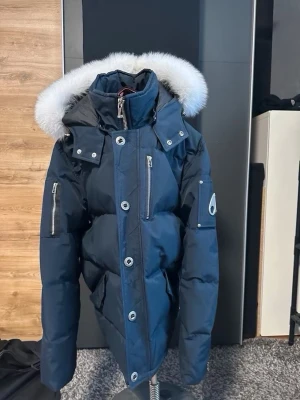 Moose Knuckles mörkblå dunjacka med päls - Snygg mörkblå dunjacka från Moose Knuckles med fluffig vit päls på huvan, stora silvriga knappar och dragkedjor. Jackan har lång ärm, flera fickor med dragkedja och ikonisk logga på ärmen. Perfekt för kalla dagar och riktigt stilren look.