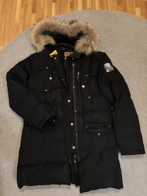 Svart vinterjacka Parajumpers - Svart Parajumpers dunjacka med fluffig päls på huvan, flera fickor med knappar och dragkedja framtill. Perfekt för kalla dagar och har en lång, rymlig passform.