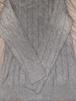 Grå stickad half zip tröja Polo Ralph Lauren - Säljer en snygg grå kabelstickad tröja från Polo Ralph Lauren med half zip och långärmad design. Tröjan har ribbade muddar och krage, samt den klassiska Polo-loggan broderad på bröstet. Perfekt för dig som gillar stilrena och tidlösa plagg.