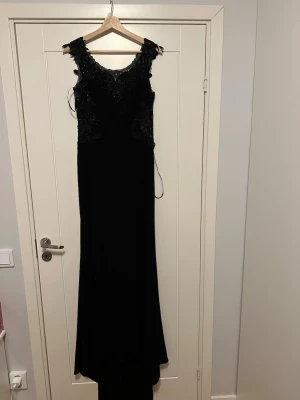 Svart långklänning med spetsdetaljer - Elegant svart aftonklänning med vacker spetsdetalj upptill. Klänningen har en figurnära passform och en lång, böljande kjol.  Använd vid ett tillfälle och därefter kemtvättad. Det anmärkningsbara är att sömmen inte har sytts ordentligt vid ena sidan på tre ställen, se bilder. Det är dock inget som stör, jag fick den hemskickad i det skicket när jag beställde den och hade inte tid att byta :) kan enkelt sys om man har symaskin eller är händig :) Lite nertrampad på ett ställe men annars inget 