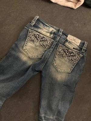 Miss Me Signature Bootcut Jeans, strl 25 - Snygga blå Miss Me jeans i bootcut-modell med coola broderade bakfickor dekorerade med pärlor och paljetter. Jeansen har slitningar och kontrastsömmar, samt detaljerade knappar. Perfekt för dig som gillar statement-plagg och vill sticka ut.