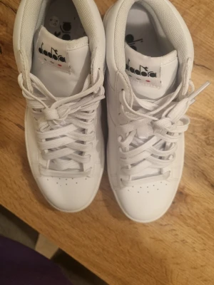 Vita Diadora sneakers high-top strl 36 - Säljer ett par fräscha vita high-top sneakers från Diadora i storlek 36. Skorna har snörning, rund tå och är tillverkade i syntetmaterial med mjuk vadderad kant. Klassisk logga på både plös och häl. Perfekta för dig som gillar stilrena och sportiga skor.