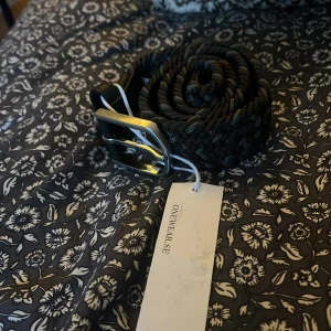 Svart och grå flätat bälte från Onewear - Snygg svart och grå flätad bälte från Onewear med stilren design och metallspänne. Bältet har en klassisk form och är tillverkad i ett syntetmaterial som ger en modern känsla. Perfekt för dig som vill ha en cool och enkel look.