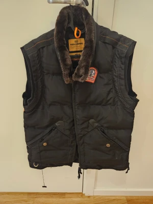 Svart pjs  - Svart dunväst med snyggt broderat märke på bröstet och mjuk fuskpäls runt kragen. Västen har två stora fickor med dragkedja och tryckknapp, samt orange detalj i nacken. Slim fit-modell, perfekt för kalla dagar. 