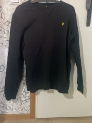 Svart tröja från Lyle & Scott, M - Svart långärmad tröja från Lyle & Scott i storlek M. Klassisk design med rund hals och gul broderad logga på bröstet. Mjuk och skön bomullskvalitet som passar perfekt till chill eller häng med kompisar.