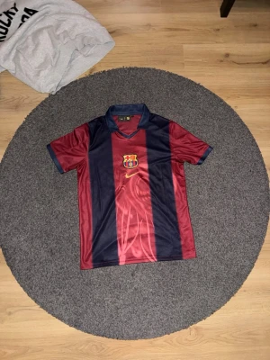 FC Barcelona x Travis Scott Fotbolls Tröja - Säljer en FC Barcelona x Travis Scott fotbollströja i storlek L. Tröjan är vinröd och marinblå med klubbmärke och Nike-logga framtill, samt 'Cactus Jack', nummer 2 och 'BARÇA' på ryggen. Klassisk krage och tillverkad i polyester.