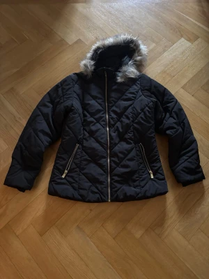 Svart pufferjacka med fuskpäls från Lindex - Svart pufferjacka från Lindex med snyggt quiltat mönster och huva med fuskpäls. Jackan har guldfärgade dragkedjor och två fickor fram.