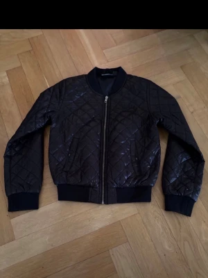 Svart quiltad bomberjacka stl 152 - Snygg svart bomberjacka med quiltat mönster och dragkedja framtill. Jackan har ribbade muddar vid ärmslut och midja, samt en klassisk rund krage. Perfekt för dig som gillar en stilren look med lite edge.