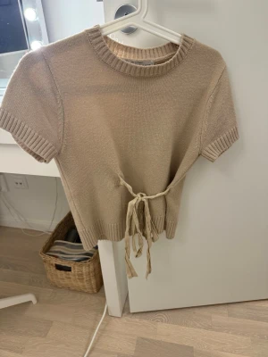 Beige stickad tröja med knyt från Zara - Säljer en beige stickad tröja från Zara i storlek S. Tröjan har korta ärmar, rund hals och ett snyggt knytband i midjan som ger en cool detalj. Ribbstickade muddar vid ärmslut och nederkant. Perfekt till jeans eller kjol för en chill look.