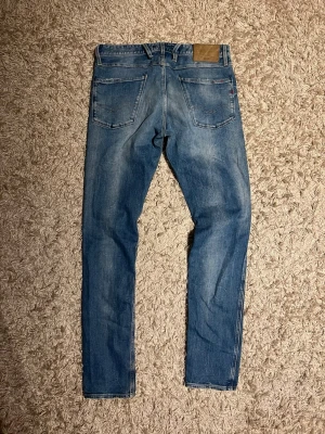 Replay anbass jeans slim fit herr w32 - Replay anbass jeans i slim passform. Storlek W32. Skön wash o små slitningar. Mycket bra skick övrigt, förutom två bälteshåller har lossnat nedifrån på baken, den i mitten fungerar, se bild. Postas samma dag som köp eller dagen därpå🙌💯