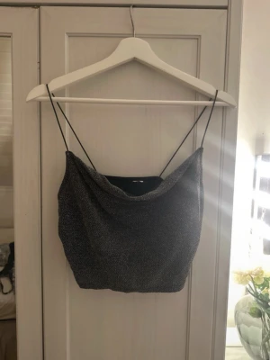 Glittrig silver croppad linnetopp - Säljer en glittrig silverfärgad croppad linnetopp med tunna svarta axelband. Toppen har en snygg draperad framsida och är perfekt för dig som vill sticka ut. Materialet är mjukt och skimrande, vilket ger en lyxig känsla.