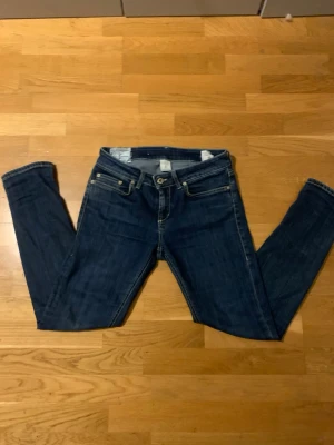 Mörkblå jeans med smal passform - Snygga mörkblå jeans med klassisk femficksdesign och smal passform. Jeansen har kontrastsömmar och metallknapp i midjan. Perfekta för dig som gillar en stilren och enkel look. Materialet är slitstarkt jeans och färgen är djup blå.