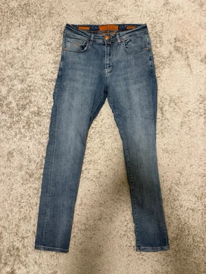 Blå jeans från Jacob Cohën Limited Edition - Säljer ett par blå jeans från Jacob Cohën Limited Edition med snygga orangea sömmar och detaljer. Klassisk femficksmodell, rak passform och normal midja. Jeansen har en broderad J på myntfickan och patch i läder bak i midjan. Perfekta till en avslappnad stil.