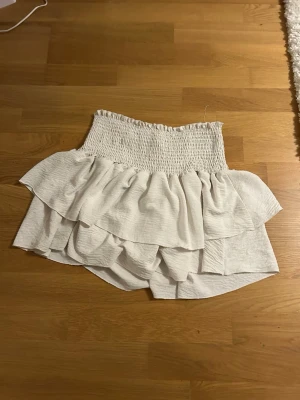 Vit volangkjol med smock - Supersöt vit kortkjol med bred smock i midjan och dubbla volanger som ger en riktigt flowig och lekfull look. Kjolen är gjord i ett lätt och skönt tyg, perfekt för varma dagar. Den har fläckar som bara syns i vissa ljus o syns därefter inte på bilden. Den är köpt second hand och säljer vidare den då den ej används.