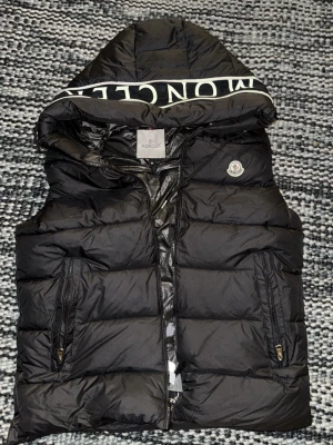Svart dunväst från Moncler med huva - Svart dunväst från Moncler med stor huva och tydlig logotyp på huvkanten. Västen har två fickor med dragkedja, quiltad design och klassisk Moncler-märke på bröstet. Materialet är glansigt och känns riktigt lyxigt, perfekt för kyliga dagar.