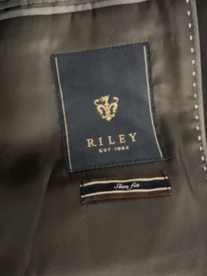 Svart slim fit kavaj från Riley - Svart kavaj från Riley i slim fit-modell med klassisk krage och två fickor med lock framtill. Snygg och stilren design som passar perfekt till skjorta. Materialet känns mjukt och har en elegant finish. Perfekt för dig som gillar en clean look.