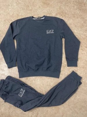 EA7 Emporio Armani mjukisbyxor grå - Snygga grå mjukisbyxor från EA7 Emporio Armani med logga tryckt på ena benet. Byxorna har resår i midjan och muddar vid bensluten för en skön och avslappnad passform. Perfekta för chill eller träning.