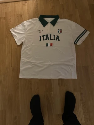 Vit Italia t-shirt med grön krage - Säljer en vit t-shirt med grön krage och gröna detaljer på ärmarna. Tryck med texten 'ITALIA' och italienska flaggan på bröstet samt ett litet emblem. Perfekt för dig som gillar sportig stil och vill visa ditt stöd för Italien.