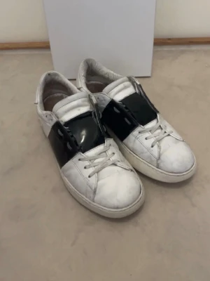 Valentino Open sneakers vit/svart - Säljer ett par Valentino Open sneakers i vitt skinn med bred svart läderdetalj över mitten. Klassisk rund tå, platt sula och snörning framtill. Snygg kontrast mellan det vita och svarta, perfekt för dig som gillar stilrena designer. Helt ok skick men ändå tecken på användning. Går alltid o pruta