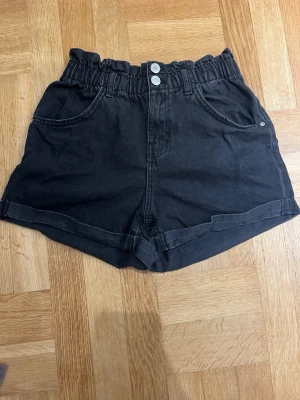 Svarta  shorts  - Säljer ett par svarta högmidjade shorts från Zara med två framfickor, två bakfickor och dubbla knappar i midjan. Shortsen har en snygg rynkad midja och är gjorda i mjuk bomull. Perfekta för chill dagar och har en loose passform.