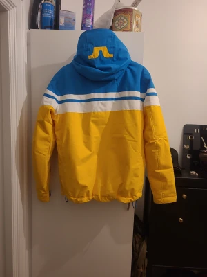 Färgglad skidjacka från J.Lindeberg - Säljer en jacka från J.Lindeberg i blått, gult och vitt med coola ränder över bröstet och huva med snörning. Jackan har dragkedja framtill, justerbara ärmslut och är tillverkad i 100% polyester. Perfekt för dig som gillar färg och vill sticka ut.