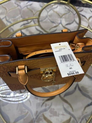 Brun handväska Michael Kors Hamilton - Snygg handväska från Michael Kors i modellen Hamilton. Väskan är brun med MK-logga och har ett reptilmönstrat parti på framsidan. Guldiga metalldetaljer, lås och nyckel hänger framtill. Väskan är tillverkad i skinn och har både handtag och axelrem samt dragkedja inuti. Väskan är köpt hos Michael kors i Canada.