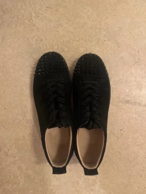 Christian Louboutin Skor - Säljer ett par svarta sneakers i mocka med coola svarta nitar på tån och klassisk röd sula. Skorna har snörning och en stilren, rund tå. Perfekta för dig som vill sticka ut med en edgy vibe.