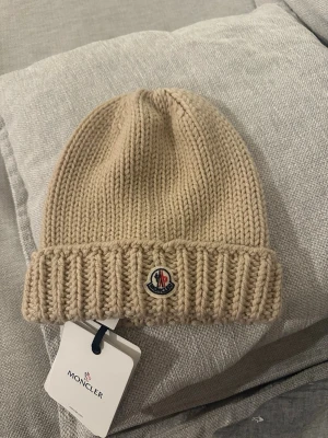 Moncler beige stickad mössa  - Snygg beige stickad mössa från Moncler med klassisk ribbad kant och broderad logotyp framtill. Mössan är tillverkad i tjockt garn som ger en skön och varm känsla. Perfekt accessoar för kyliga dagar och enkel att matcha med din vinterstil.